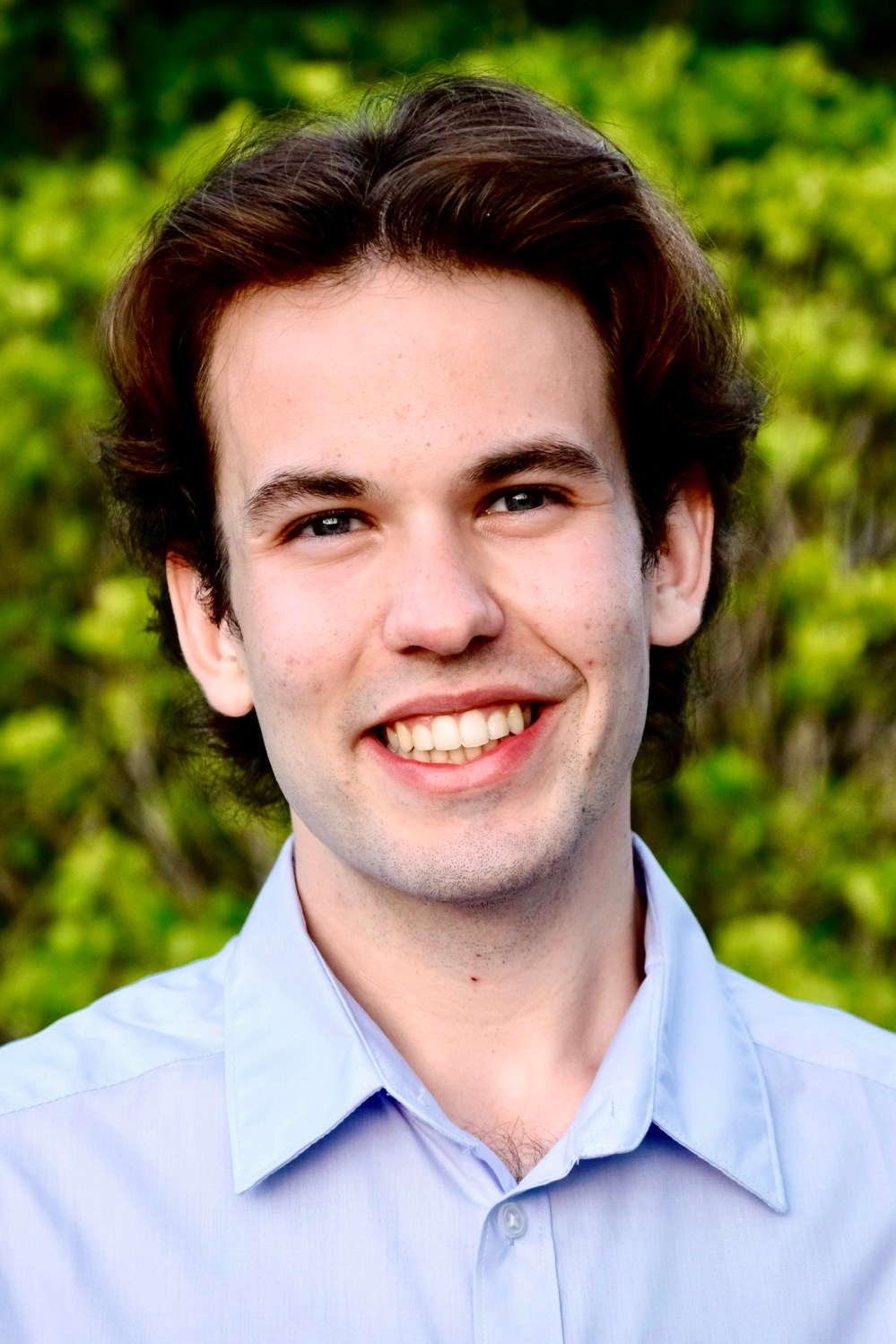 Young male smiling, wearing a light blue shirt, with green foliage in the background.
