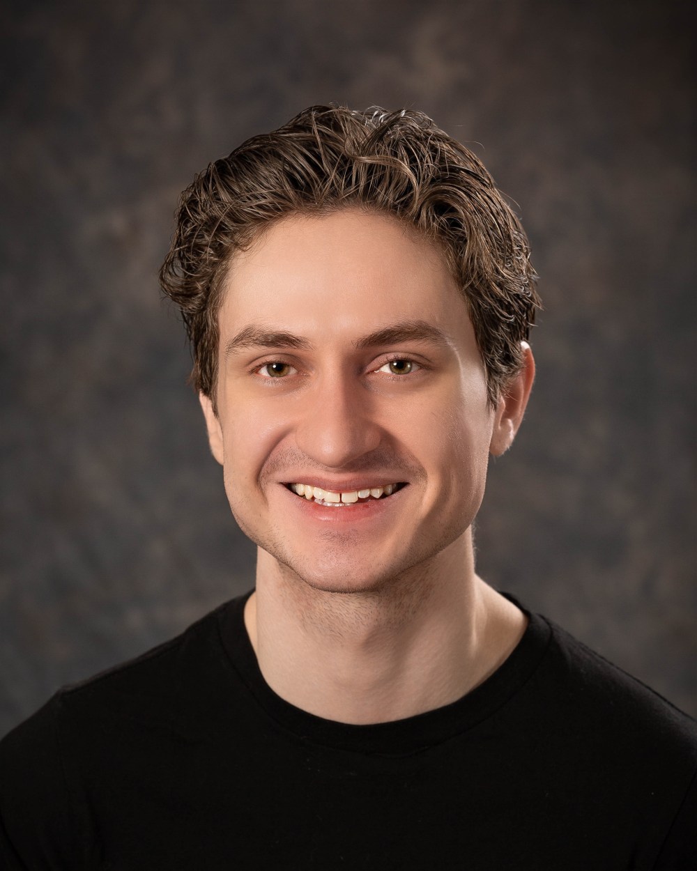 Smiling person with short, wavy hair, wearing a black shirt, against a dark textured background.