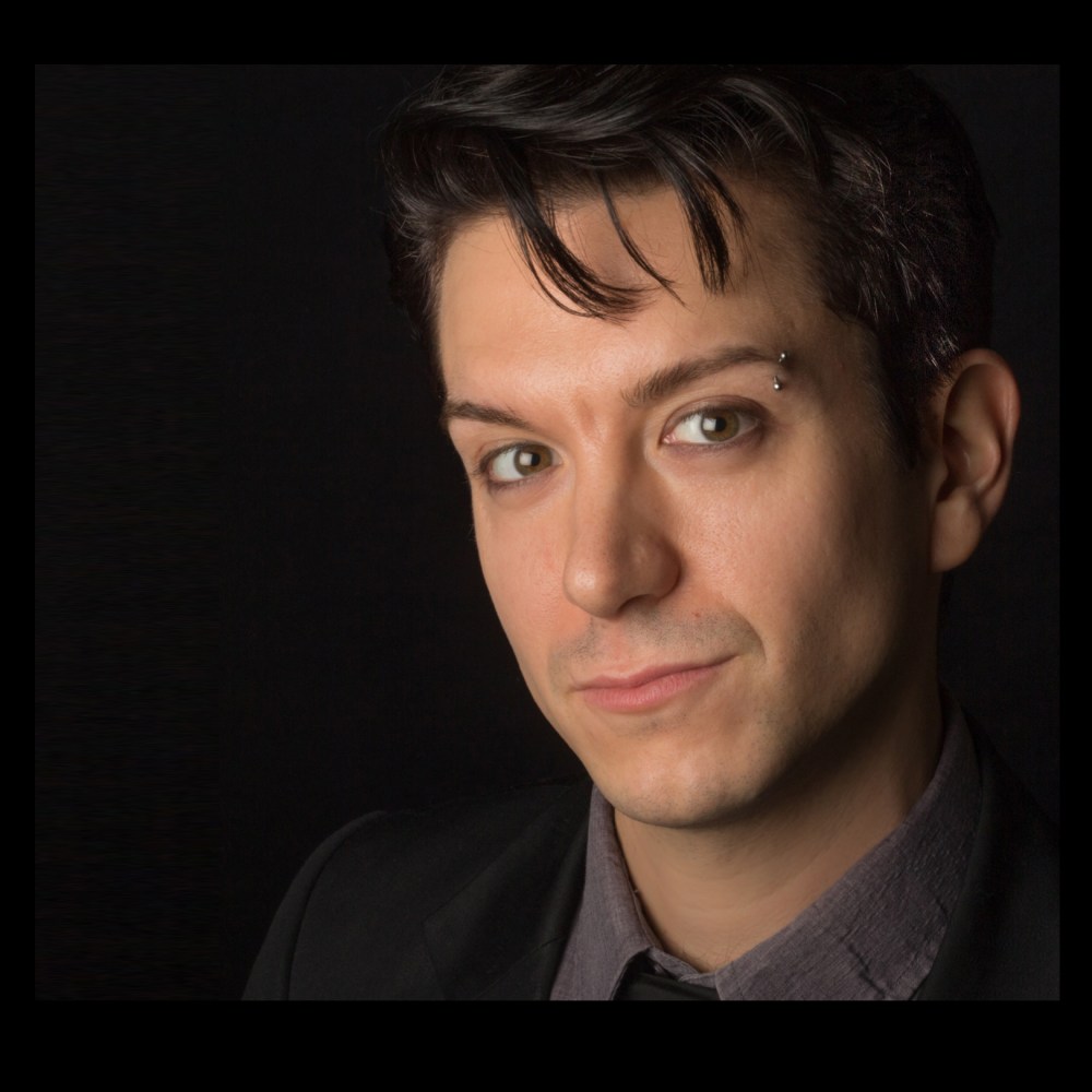 Young man with dark hair and eyebrow piercing, wearing a suit jacket and shirt, against a dark background.