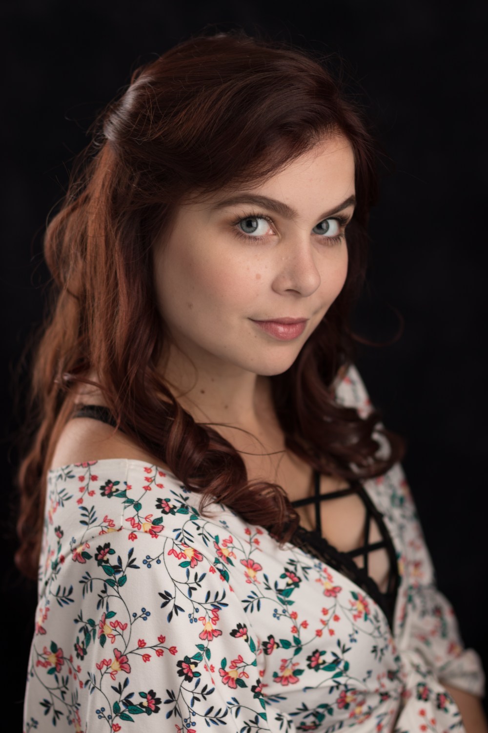 Woman with long auburn hair, wearing floral top, poses against dark background.