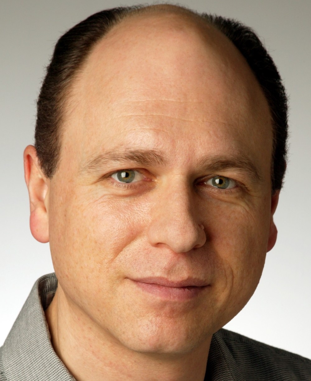 Close-up of a man with short dark hair, wearing a gray shirt, and looking at the camera.